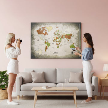 Two women photographing and pinning to a large sepia canvas world map above a gray sofa in a pink room
