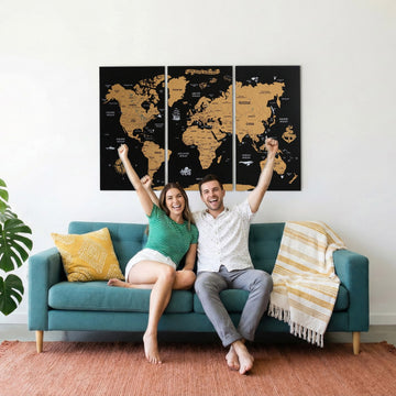 Black cork push pin world map in three panels above a teal sofa, with a smiling couple cheering