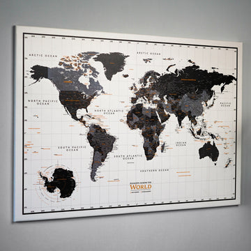 Framed black and gray canvas world map hung on a gray wall, seen from a side angle