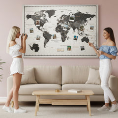 Black and gray canvas world map with photo pins above a sofa, two women looking at the map in a living room