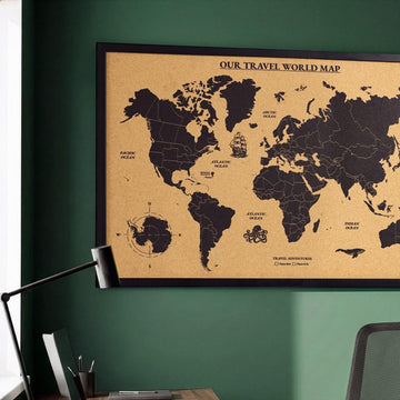 Close-up of the framed cork world map with black continents, the title band and Antarctica beside a desk lamp