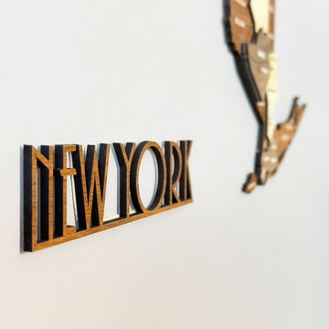 Side view of the NEW YORK cut-out lettering beside the edge of the layered wooden New York map on a gray wall
