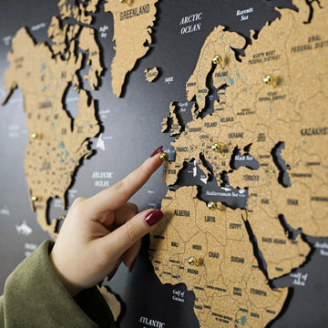 Black cork world map with Africa and Greenland visible, a hand placing a gold pin on Europe