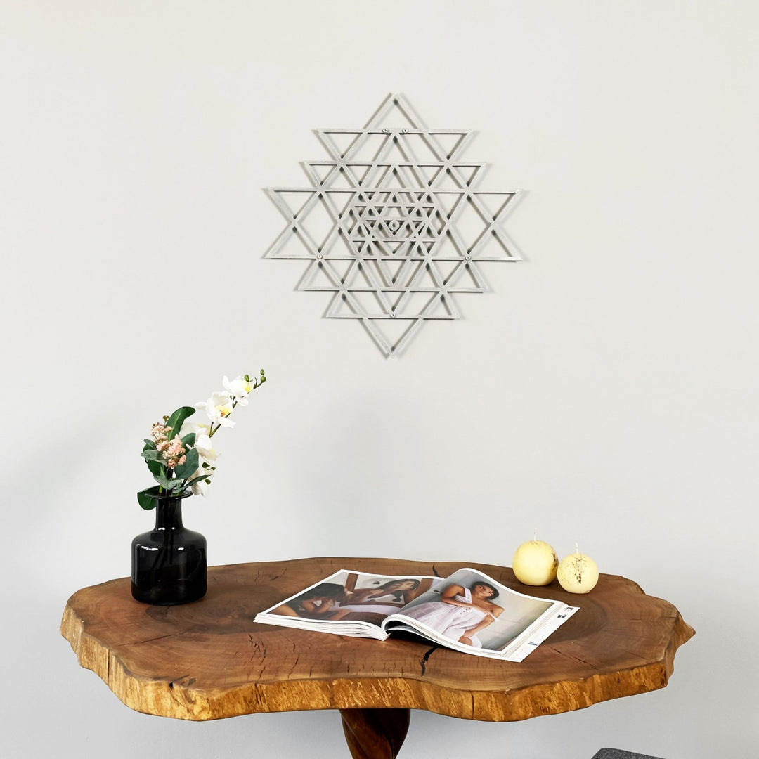 Wooden table with a vase, flowers, and magazines against a white wall with a silver sri yantra scared triangle wall art piece.