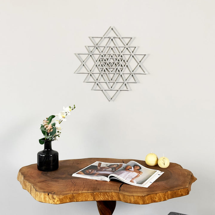 Wooden table with a vase, flowers, and magazines against a white wall with a silver sri yantra scared triangle wall art piece.