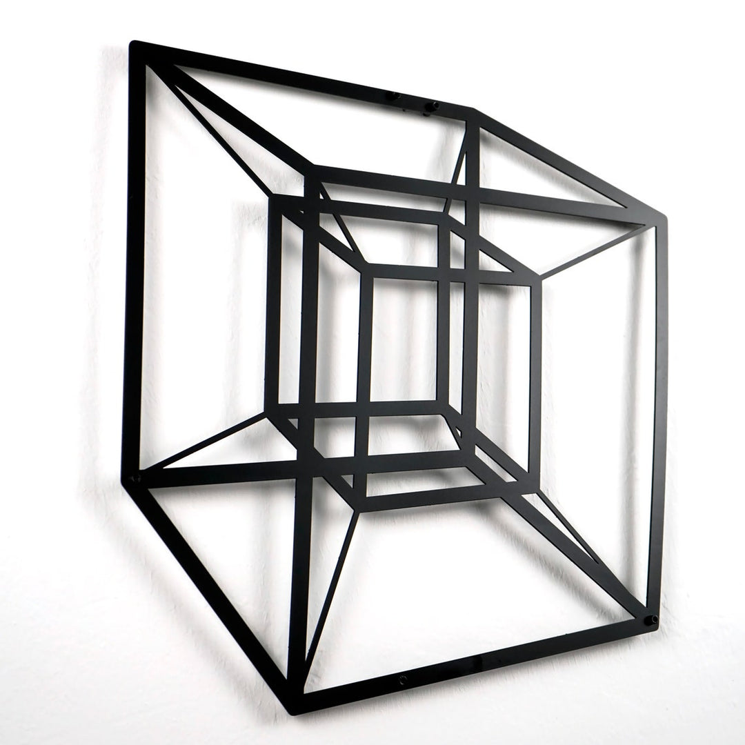 Black tesseract cube metal wall art on a white wall, close view