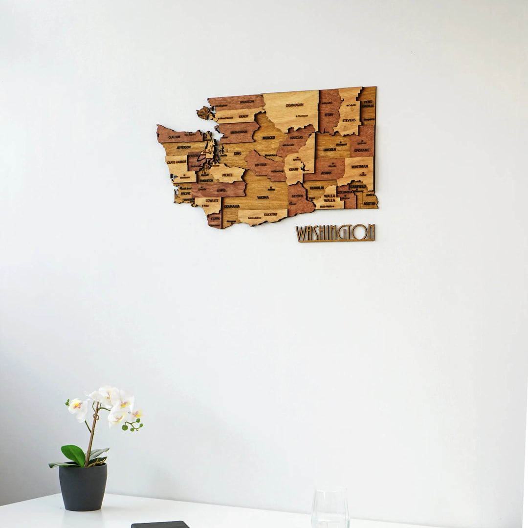 Washington State wooden map centered on a white wall, with an orchid and desk edge below