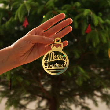 Gold mirror acrylic Merry Christmas bauble ornament held up in front of a Christmas tree